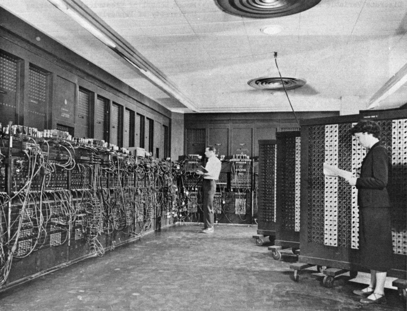 Black and white photo of two programmers working at ENIAC's wall of panels and cables